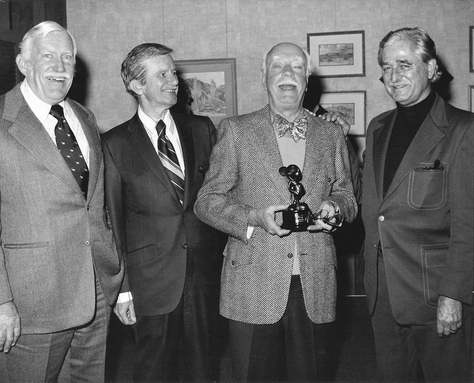 Four smiling older men in suits stand together indoors. One man in the center holds a small statue or award. Framed pictures hang on the wall behind them.